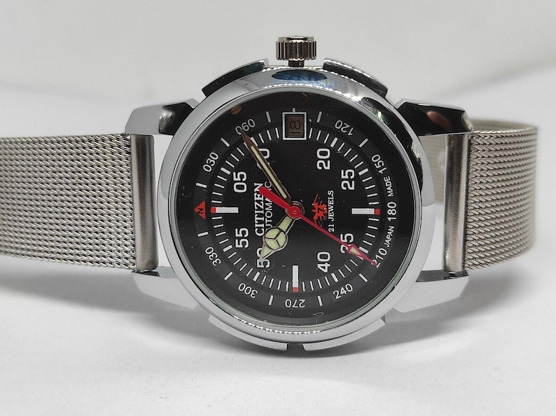 May include: A silver-toned Citizen automatic watch with a black dial, featuring white and red markings. The watch has a mesh band and a date display at the 3 o'clock position. The watch face displays the text "CITIZEN AUTOMATIC 21 JEWELS MADE IN JAPAN".