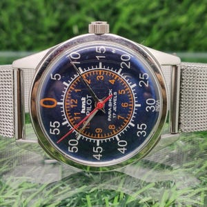 May include: Silver wristwatch with a dark blue face, orange and white numerals and hands. The watch face displays "hmt PILOT" and "PARA SHOCK 17 JEWELS". The watch has a silver mesh strap.