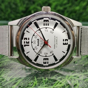 May include: A silver wristwatch with a white face and black numerals. The watch has a mesh strap and a red second hand. The watch face includes the text "PARA SHOCK 17 JEWELS" and "PILOT".