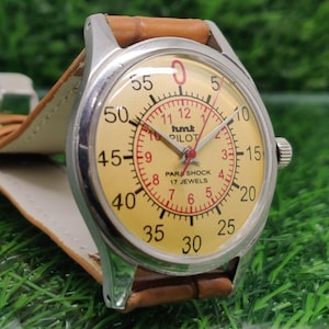 May include: A vintage wristwatch with a light yellow face, featuring black and red numerals and hands. The watch has a silver-colored case and a brown leather strap. The watch face reads "hmt PILOT PARASHOCK 17 JEWELS".