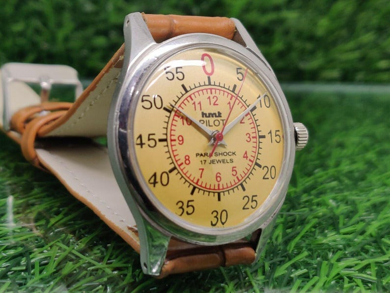 May include: A vintage wristwatch with a light yellow face, featuring black and red numerals and hands. The watch has a silver-colored case and a brown leather strap. The watch face reads "hmt PILOT PARASHOCK 17 JEWELS".