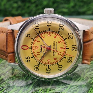 Może przedstawiać: Zegarek vintage z żółtą tarczą, czarnymi cyframi i czerwonymi akcentami. Zegarek ma brązowy skórzany pasek i srebrną kopertę. Na tarczy widnieje napis "hmt PILOT PAR SHOCK 17 JEWELS".