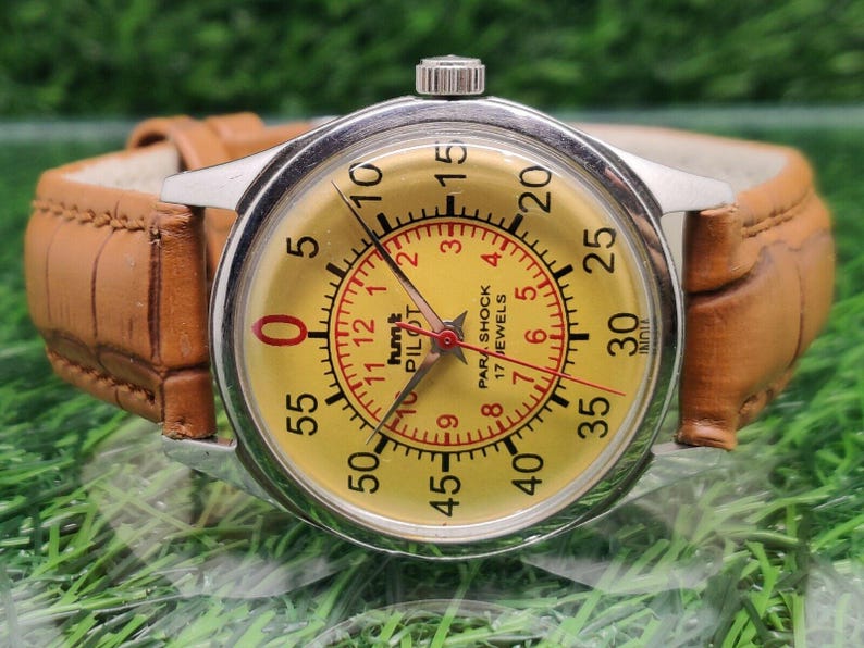 May include: A vintage wristwatch with a yellow face, black numerals, and red accents. The watch has a brown leather band and a silver-colored case. The watch face includes the text "hmt PILOT PAR SHOCK 17 JEWELS".
