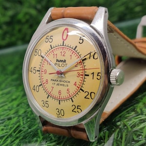 May include: Vintage HMT Pilot watch with a cream-colored face, red numerals, and a brown leather strap. The watch has a silver-colored case and features the text "hmt PILOT PARA SHOCK 17 JEWELS".