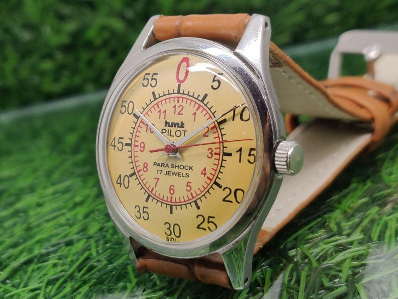 May include: Vintage HMT Pilot watch with a cream-colored face, red numerals, and a brown leather strap. The watch has a silver-colored case and features the text "hmt PILOT PARA SHOCK 17 JEWELS".
