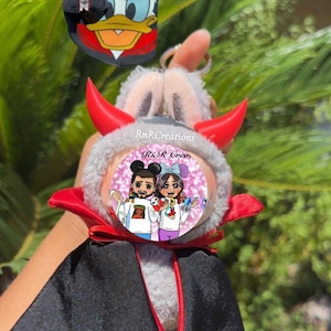 May include: A plush devil-themed keychain with red horns, a black cape, and a gray fur body. The keychain has a cartoon image of two people on the front. A small black bag with a Donald Duck design is also visible.