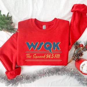 May include: A red sweatshirt with the text "WSQK The Squawk 94.5 FM" in blue and gold. The sweatshirt is folded with the sleeves and hood visible. Decorative elements include a small Christmas tree, pinecones, and a red mug.