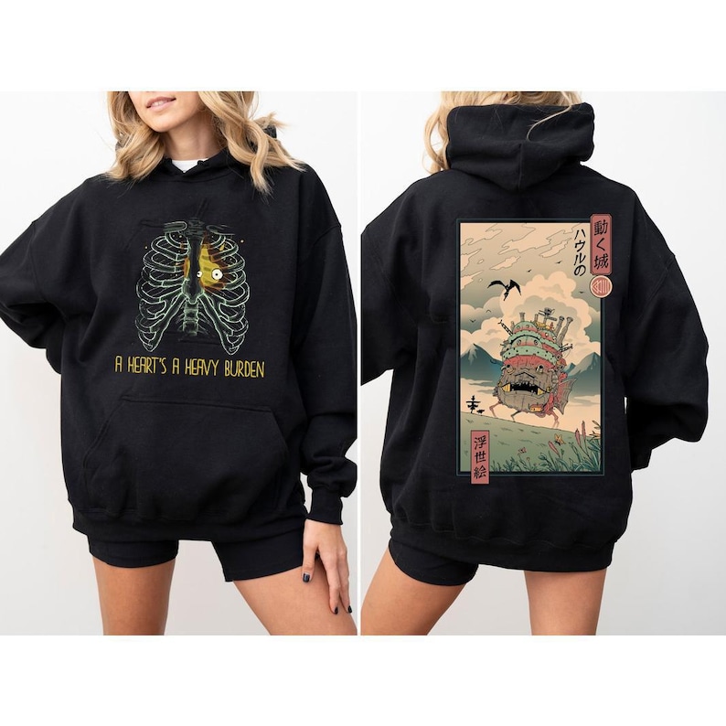 Puede incluir: Sudadera con capucha negra con un gr&aacute;fico frontal de una caja tor&aacute;cica con un coraz&oacute;n amarillo y el texto "A HEART'S A HEAVY BURDEN." La parte trasera presenta una ilustraci&oacute;n colorida de un castillo en movimiento con texto japon&eacute;s y un paisaje monta&ntilde;oso.