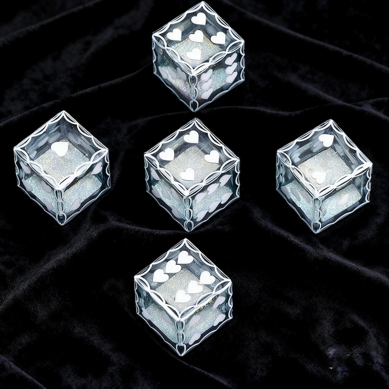 May include: Five transparent cube-shaped boxes featuring white heart-shaped patterns on the edges and sides. Each box contains a shimmering, light-reflecting silver material inside, visible through the clear walls of the containers.