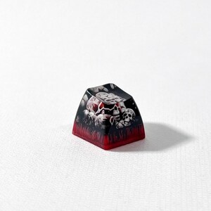 Puede incluir: Una tecla personalizada para teclado mec&aacute;nico con un gr&aacute;fico detallado de varias calaveras en tonos blancos y grises sobre un fondo oscuro, con una base roja transl&uacute;cida que se transforma en un color negro intenso en la superficie superior.