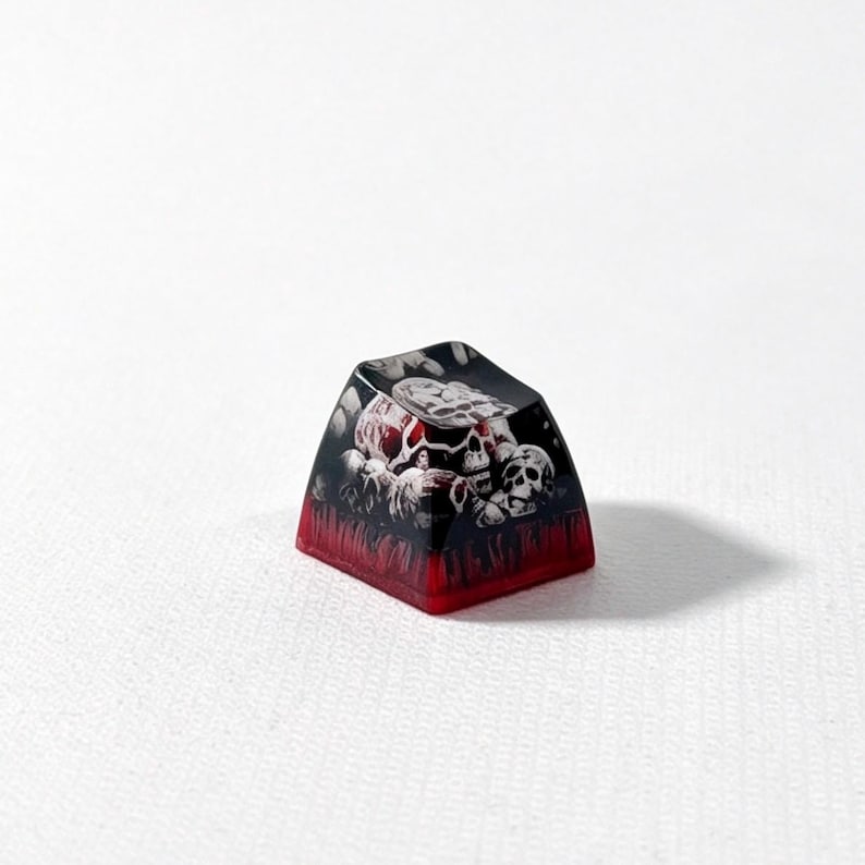 Puede incluir: Una tecla personalizada para teclado mec&aacute;nico con un gr&aacute;fico detallado de varias calaveras en tonos blancos y grises sobre un fondo oscuro, con una base roja transl&uacute;cida que se transforma en un color negro intenso en la superficie superior.