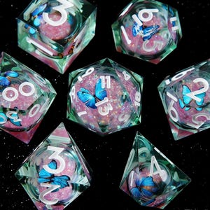May include: Set of seven polyhedral dice with a butterfly theme. The dice are clear with a green tint, filled with pink glitter, and feature blue butterfly accents. White numbers are visible on each face, suitable for tabletop games.