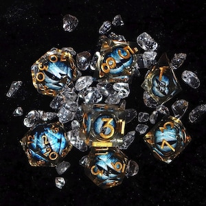 May include: Set of seven polyhedral dice with a blue and white eye design, gold numbering, and gold trim. The dice are surrounded by clear, faceted crystals on a black surface. A unique set for tabletop gaming.