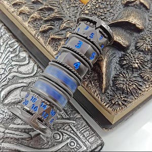 May include: A metallic, multi-sided dice roller with blue numbering and accents. The dice roller is cylindrical with multiple rotating sections. The background features a detailed, textured surface with a fantasy theme.