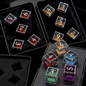 May include: A collection of multi-colored translucent dice with white numbers and intricate silver-toned filigree edges. The dice are arranged in black velvet-lined metal tins, featuring various colors including red, orange, blue, green, and purple.
