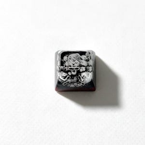 Puede incluir: Una tecla mec&aacute;nica cuadrada para teclado con un gr&aacute;fico detallado en blanco y negro de varias calaveras apiladas. La tecla tiene un acabado brillante con una base oscura y transl&uacute;cida que se transforma en un tono rojo intenso en el borde inferior.
