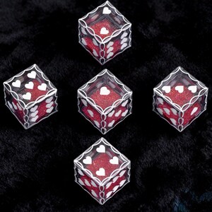 May include: Five transparent cubic dice featuring white heart-shaped pips on each face. The interior of each die is filled with a vibrant red glitter material, and the edges are outlined with a decorative white filigree pattern.