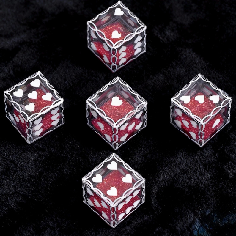 May include: Five transparent cubic dice featuring white heart-shaped pips on each face. The interior of each die is filled with a vibrant red glitter material, and the edges are outlined with a decorative white filigree pattern.