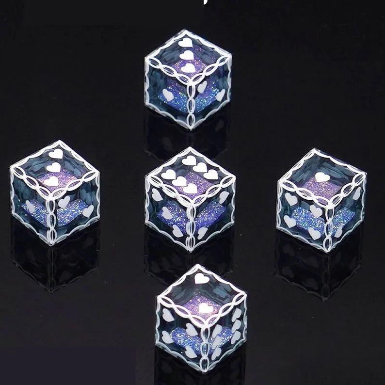 May include: A set of five translucent blue dice with glittery purple interiors. Each die features white heart-shaped pips on the faces and a white filigree-style border pattern along the edges, creating a decorative look for these gaming pieces.