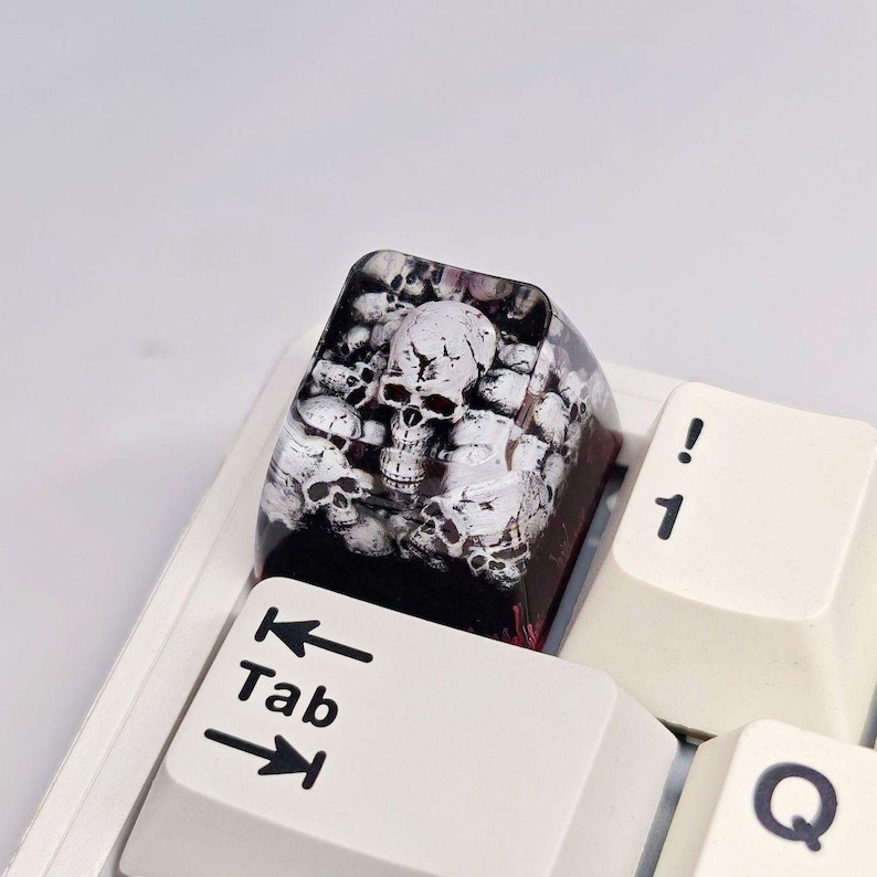 Puede incluir: Una tecla mec&aacute;nica personalizada de resina con un dise&ntilde;o detallado de m&uacute;ltiples capas de calaveras blancas incrustadas en un material transparente de tono oscuro. La tecla est&aacute; colocada en un teclado blanco junto a la tecla Tab y el 1.