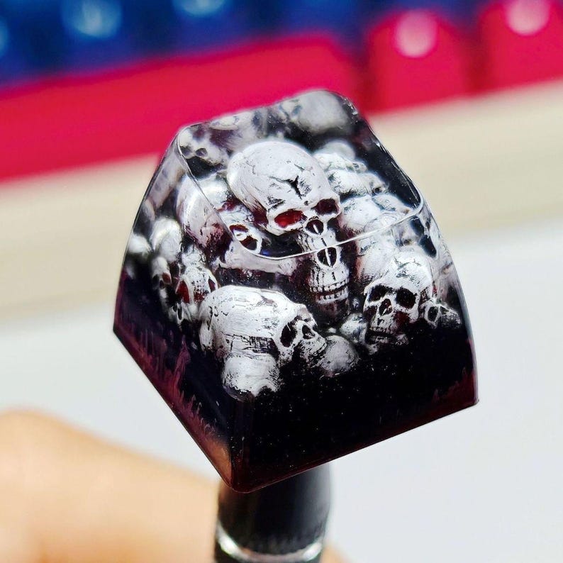 Puede incluir: Una tecla mec&aacute;nica cuadrada hecha de resina transparente con una base oscura. El interior presenta un dise&ntilde;o detallado y multicapa de varias calaveras blancas con sombreado negro y detalles en rojo, creando un efecto tridimensional.