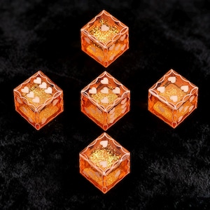 May include: A set of five translucent orange dice with gold glitter suspended inside. Each die features white heart-shaped pips on the faces and a decorative white filigree border framing the edges of each side of the cube-shaped game pieces.