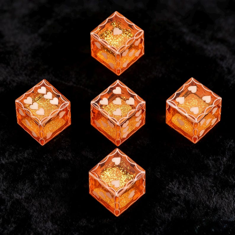 May include: A set of five translucent orange dice with gold glitter suspended inside. Each die features white heart-shaped pips on the faces and a decorative white filigree border framing the edges of each side of the cube-shaped game pieces.