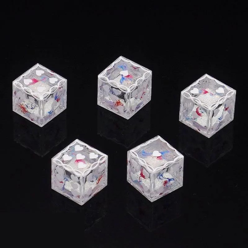 May include: Five clear, cube-shaped dice featuring white heart icons on each face. The interior of each die contains a glittery, textured substance with visible red and blue accents, while the edges are outlined with a white, decorative filigree pattern.
