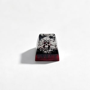 Puede incluir: Una tecla para teclado mec&aacute;nico con un gr&aacute;fico detallado de varias calaveras en tonos blanco, gris y negro. La base de la tecla presenta un color rojo transl&uacute;cido profundo con un patr&oacute;n oscuro similar a llamas en el borde inferior.