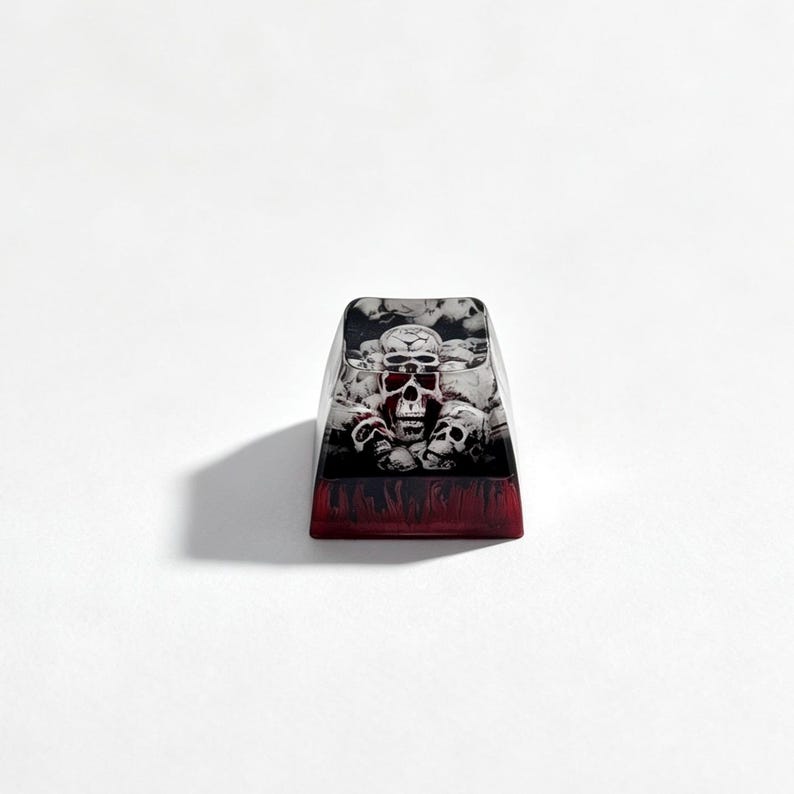 Puede incluir: Una tecla para teclado mec&aacute;nico con un gr&aacute;fico detallado de varias calaveras en tonos blanco, gris y negro. La base de la tecla presenta un color rojo transl&uacute;cido profundo con un patr&oacute;n oscuro similar a llamas en el borde inferior.