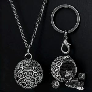 Puede incluir: Un conjunto de collar con colgante y llavero de color plateado. El colgante es una esfera con números, y el llavero incluye un recipiente en forma de dado con dados en su interior. El conjunto está diseñado para los entusiastas de los juegos de mesa.