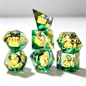 May include: A set of polyhedral dice made of translucent green resin, each containing a miniature yellow rubber duck. The dice feature gold-colored numbers etched into the surfaces. The set includes various shapes, including d20, d12, d10, d8, and d4, stacked in three distinct columns.