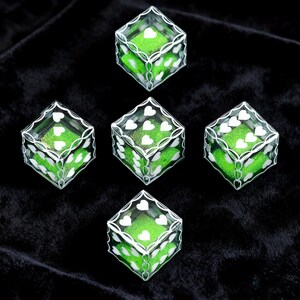 May include: A set of five transparent dice with white heart-shaped pips and a bright green glittery interior. Each die features a white filigree-style border pattern along the edges, creating a decorative frame for the heart symbols on each face.