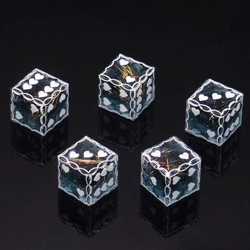 May include: A set of five translucent blue dice with white heart-shaped pips and white chain-link borders. Each die contains embedded iridescent gold-toned metallic fibers visible through the clear resin material, creating a reflective effect.