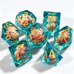 May include: A set of seven polyhedral dice made of clear teal resin, each containing a miniature orange basketball inside. The dice feature gold-colored numbers etched into the surfaces, including a d20, d12, d10, d8, d6, and d4, arranged in a cluster.