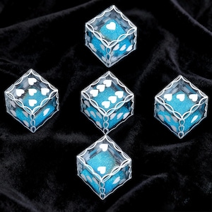 May include: Five clear, cube-shaped containers with white heart-shaped patterns printed on the edges and corners. Each container is filled with bright blue, glittery material, creating a vibrant contrast against the white heart designs.