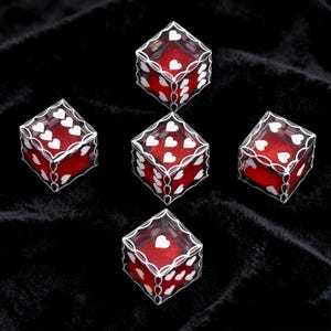 May include: Five six-sided dice featuring a clear outer shell with white heart-shaped pips and a red glittery interior. Each die displays a different number of white hearts on its faces, arranged in a standard dice configuration on a black surface.