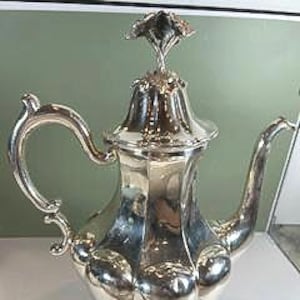 Puede incluir: Una tetera plateada con un adorno floral decorativo y un asa ornamentada. La tetera tiene un cuerpo bulboso y se apoya en tres pequeños pies. El pico se curva elegantemente y la superficie pulida refleja la luz.