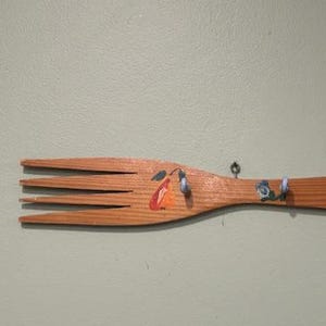 May include: A wooden fork-shaped wall hook with multiple small hooks. The wooden fork is painted with colorful floral designs and has several small hooks for hanging items. The background is a solid light green color.