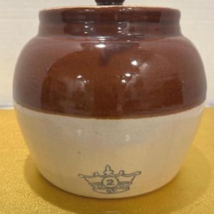 Puede incluir: Un tarro de cerámica de dos tonos con tapa. La mitad superior es marrón brillante, mientras que la mitad inferior es blanquecina. Un emblema de corona con el número 2 está impreso en la parte inferior. El tarro es probablemente para almacenamiento en la cocina.