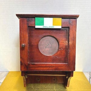 Puede incluir: Un mueble de madera marrón oscuro con una puerta pequeña y una abertura redonda. Tiene un estante en la parte inferior y un letrero con la bandera irlandesa y la palabra "IRELAND".