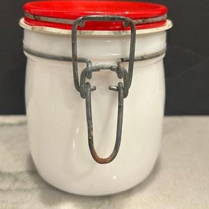 Puede incluir: Un tarro de cerámica blanco con tapa roja y cierre metálico. El tarro tiene forma redondeada y cierre de alambre metálico. La tapa es roja y el tarro es blanco. El tarro es para almacenamiento.