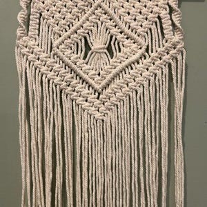 May include: A cream-colored macrame wall hanging featuring intricate knotted patterns, including a central diamond shape with a gathered tassel detail. The textile is suspended from a smooth, light-toned wooden dowel with a matching cord for hanging.