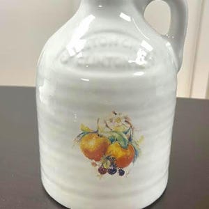May include: White ceramic jug with a handle, featuring a fruit illustration of oranges and berries. The jug has embossed text near the top. A classic kitchen or home decor item.