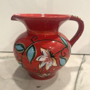 May include: A bright red ceramic pitcher featuring a hand-painted floral design with light blue leaves and white petals outlined in black. The vessel has a wide, rounded body, a flared rim with a pouring spout, and a curved handle on the side.