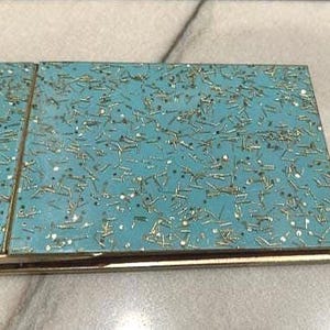 May include: A rectangular, turquoise compact with a gold-colored frame. The turquoise surface is covered in small, gold-colored, and silver-colored embellishments. The compact is closed and appears to be made of metal.