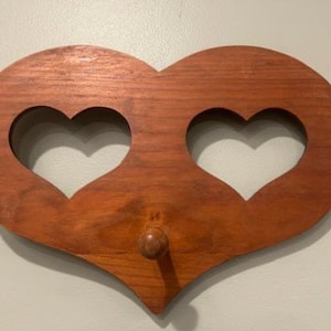 May include: A heart-shaped wooden wall hook featuring a smooth, medium-brown finish. The piece is designed with two symmetrical heart-shaped cutouts positioned side-by-side in the upper section and a single rounded wooden peg centered below.