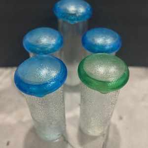 Op de afbeelding: Vijf glazen flesstoppers met getextureerde zijkanten en kleurrijke toppen. De stoppers hebben blauwe en groene toppen. Het glas heeft een getextureerd uiterlijk. De stoppers zijn in een verticale compositie gerangschikt.