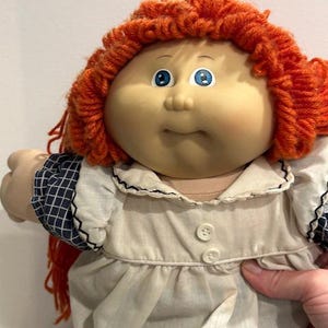 May include: A vintage doll with a round face, blue eyes, and bright orange yarn hair. The doll wears a cream-colored dress with a ruffled collar and sleeves with a blue and white checkered pattern. The doll's dress has two small buttons.