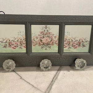 May include: A gray, distressed-finish wall-mounted organizer with three clear glass knobs. The organizer features three rectangular sections with floral artwork in pink and green tones, perfect for a bedroom or entryway.